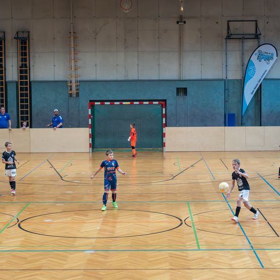 Eine Gruppe von Kindern spielt Fußball auf einem Hallenplatz. Ein Junge in Blau und Weiß tritt den Ball. Einige Leute beobachten von den Seiten.