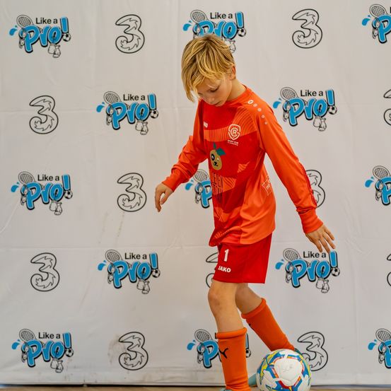 Ein junger Junge, der ein orangefarbenes Fußballtrikot trägt, übt das Kicken eines Fußballs vor einer Wand mit verschiedenen Sportlogos.