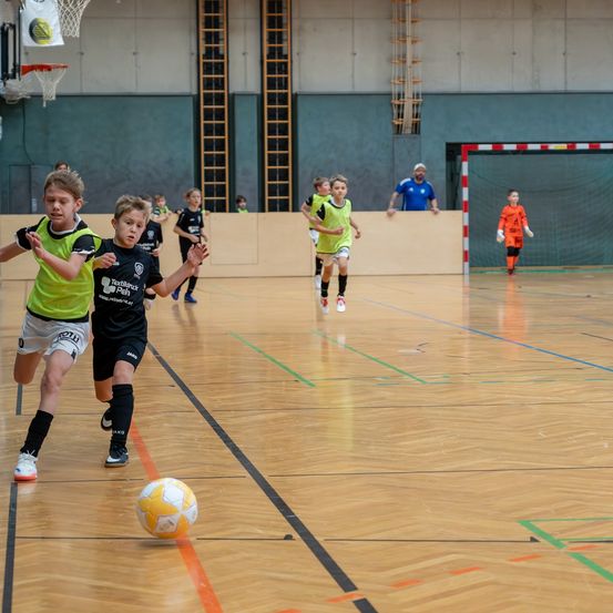 Kinder spielen Fußball auf einem Hallenplatz. Ein Junge jagt dem Ball nach, während ein anderer ihn verteidigt. Dahinter beobachtet ein Mann von den Seitenlinien.