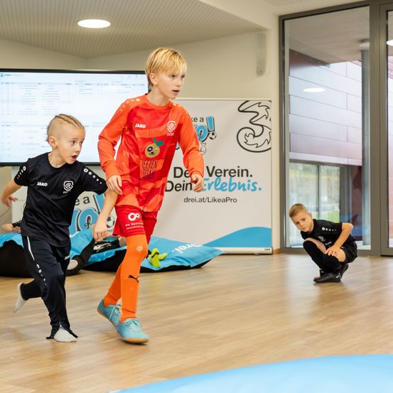 Drei Jungen in Fußballtrikots spielen auf einem Holzboden. Zwei Jungen rennen, und einer kniet. Dahinter befindet sich ein Plakat mit blau-weißem Design.