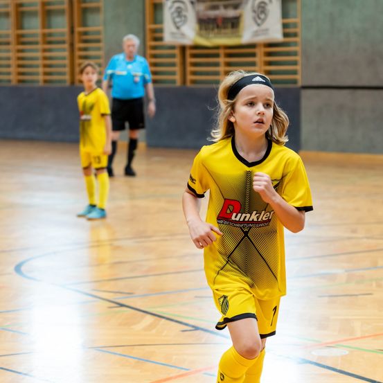 Ein junges Mädchen in einem gelben Trikot läuft auf einem Spielfeld, während ein anderes Kind und ein Schiedsrichter hinter ihr stehen. Das Spielfeld hat gelbe Markierungen und eine Holzwand mit Bannern.