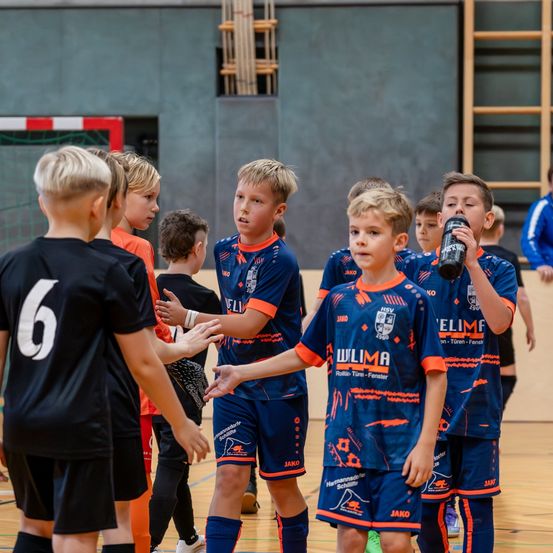 Mehrere Kinder in Sportuniformen stehen in einer Turnhalle. Sie unterhalten sich und schauen nach vorne. Ein Junge mit der Nummer 6 trinkt aus einer Flasche.