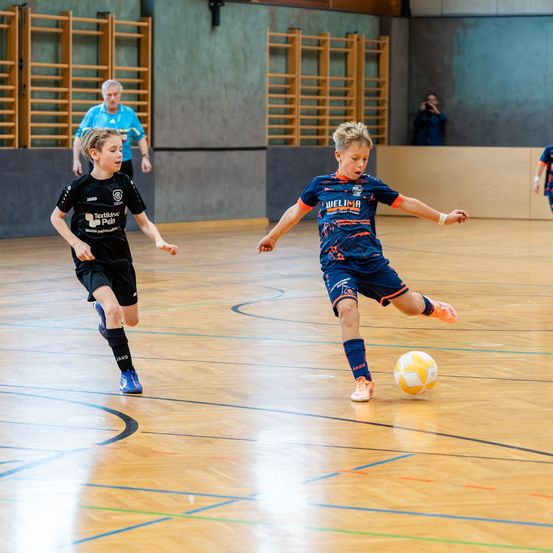 Zwei Jungen spielen Fußball auf einem Indoor-Feld. Ein Junge in einem blauen Trikot ist kurz davor, den Ball zu schießen, während der andere in Schwarz darauf zuläuft. Hinter ihnen steht ein Mann in der Nähe von Holzregalen.