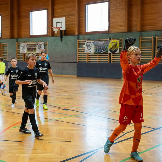 Eine Gruppe von Kindern spielt Fußball in einer Halle. Ein Junge in einem orangefarbenen Trikot hebt seine Hände, während andere spielen. Hinter ihm ist ein Basketballkorb an der Wand befestigt.