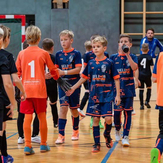 Eine Gruppe junger Jungen in orange und blauen Trikots läuft über einen Platz, wobei ein Junge aus einer Flasche trinkt.