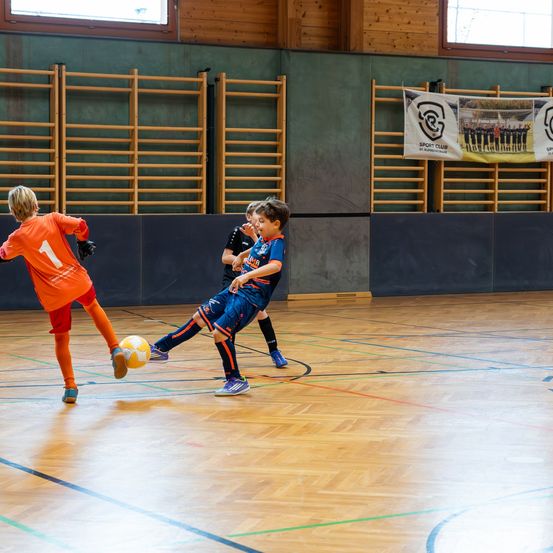 Drei Kinder spielen Hallenfußball, eines in Orange tritt den Ball, ein anderes in Blau versucht zu blocken und ein drittes in Schwarz beobachtet.