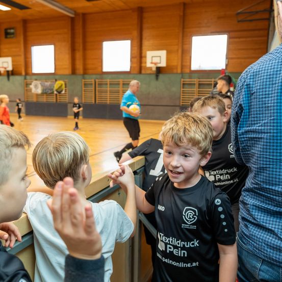 Eine Gruppe junger Jungen in einer Turnhalle, einer trägt ein schwarzes Shirt mit 'Textildruck Pein', jubelt während ein Mann mit dem Ball spielt.