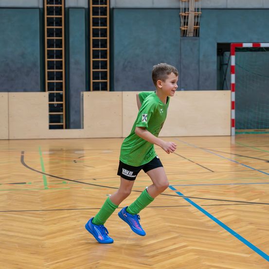 Ein junger Junge in einem grünen Fußballtrikot läuft auf einem Holzplatz mit einem Netz im Hintergrund.