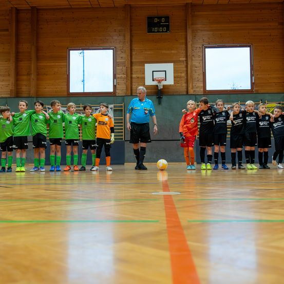 Eine Gruppe junger Fußballspieler in grünen und schwarzen Trikots steht in einer Reihe auf einem Spielfeld. Ein Erwachsener in einem blauen Hemd steht neben einem Fußball. An der Wand befindet sich ein Basketballkorb.