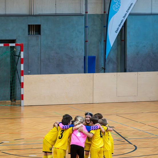 Eine Gruppe junger Spieler in gelben und rosa Trikots schmiegt sich zusammen, lächelt und lacht auf einem Hallenplatz.