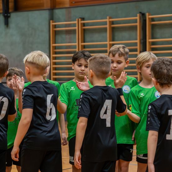 Eine Gruppe junger Jungen in Sporttrikots steht in einem Huddle, klatscht in die Hände und sieht einander an. Sie befinden sich in einer Turnhalle.