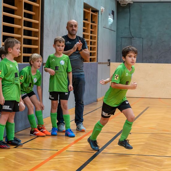 In einer Turnhalle läuft ein junger Junge in Grün, während andere zusehen. Ein Mann in Grau steht hinter ihnen. Holzregale sind an der Wand.