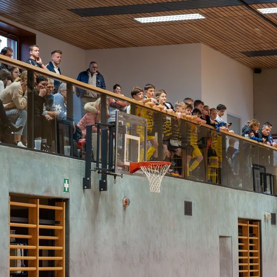Zuschauer bei einem Basketballspiel, beobachten die Spieler auf dem Spielfeld durch eine Glasbarriere, einige stehen und lehnen sich über die Barriere, andere sitzen darunter.