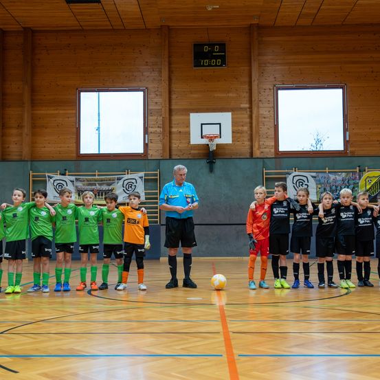 Eine Gruppe von Kindern in Fußballtrikots steht auf einem Spielfeld mit einem Ball. Ein erwachsener Mann steht unter ihnen. Das Spielfeld hat Holzwände mit Fenstern und einem Basketballkorb.