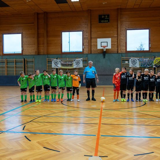 Eine Gruppe von Kindern in grünen und orangefarbenen Trikots steht in einer Turnhalle mit einem Basketballfeld, ein Mann in einem blauen Hemd und schwarzen Shorts steht in der Mitte.