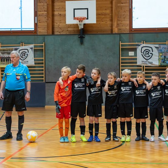 Eine Gruppe junger Fußballspieler in schwarzen Trikots steht in einer Reihe in einer Turnhalle mit einem Trainer vor ihnen. Der Boden hat einen Basketballkorb.
