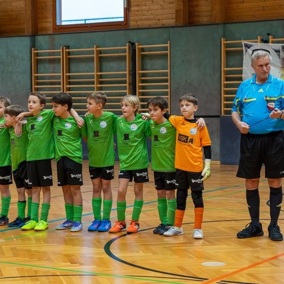 Eine Gruppe junger Jungen in grünen und orangefarbenen Fußballtrikots steht auf einem Spielfeld, während ein Trainer in der Nähe sie beobachtet.