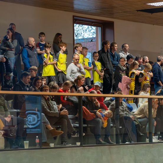 Eine Menschenmenge sitzt und steht in einer Sportarena und schaut sich ein Spiel an. Einige tragen Trikots, andere lässige Kleidung. Eine Glasbarriere trennt die Zuschauer vom Spielfeld. Ein Banner befindet sich an der Barriere.