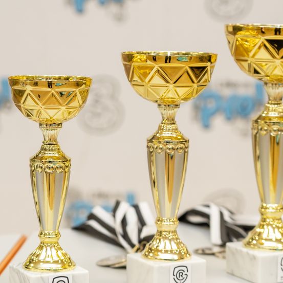 Drei goldene Pokale mit aufwendigen Verzierungen stehen auf Marmorsockeln und reflektieren Licht. Dahinter ist ein weißer Hintergrund mit einem blauen Logo zu sehen.