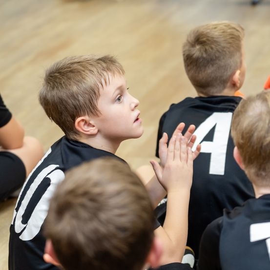 Mehrere junge Jungen in schwarzen Trikots mit Nummern sitzen auf dem Boden. Ein Junge schaut auf, während andere sich die Hand geben.