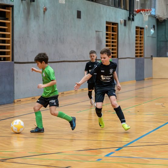 Drei Jungen spielen Fußball auf einem Hallenplatz. Zwei tragen Grün und einer trägt Schwarz. Der Junge in Schwarz jagt den Ball.