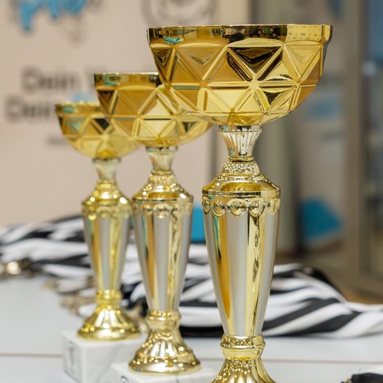 Drei goldene Pokale sind auf einer weißen Oberfläche ausgestellt, mit einem schwarz-weiß gestreiften Tuch im Hintergrund. Die Pokale haben komplizierte Designs und sind in verschiedenen Größen.