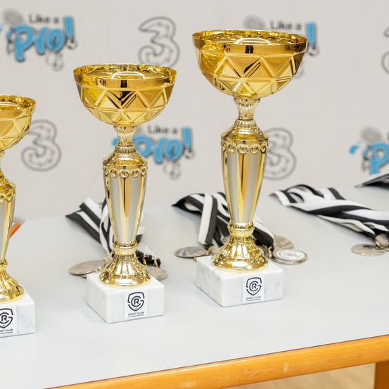Drei goldene Pokale sind auf einem weißen Tisch ausgestellt, jeder mit einem Logo und Text. Hinter den Pokalen befinden sich zwei schwarz-weiße Bänder. Der Hintergrund ist weiß mit Text und Zahlen.