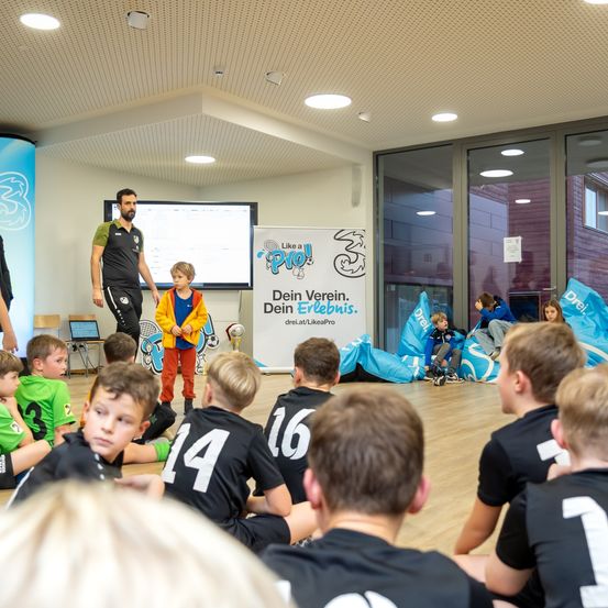 Sporthalle mit Kindern in Trikots, einer stehend, einer sitzend, Bannern und einer Projektionsleinwand.
