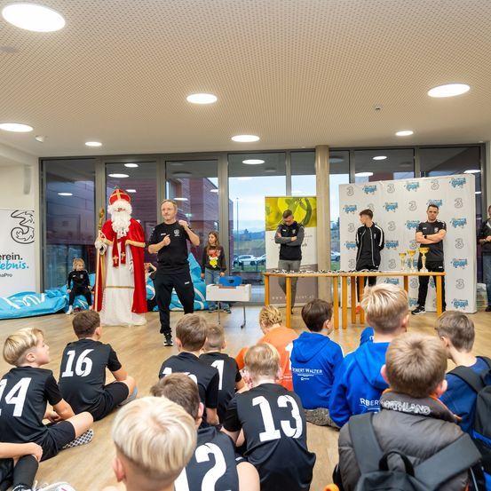 In einem Raum sitzen eine Gruppe von Kindern mit Nummern auf den Trikots auf dem Boden. Zwei Männer, einer als Weihnachtsmann gekleidet, stehen hinter einem Tisch. Der Raum hat große Fenster, Poster und eine Decke mit Lichtern.