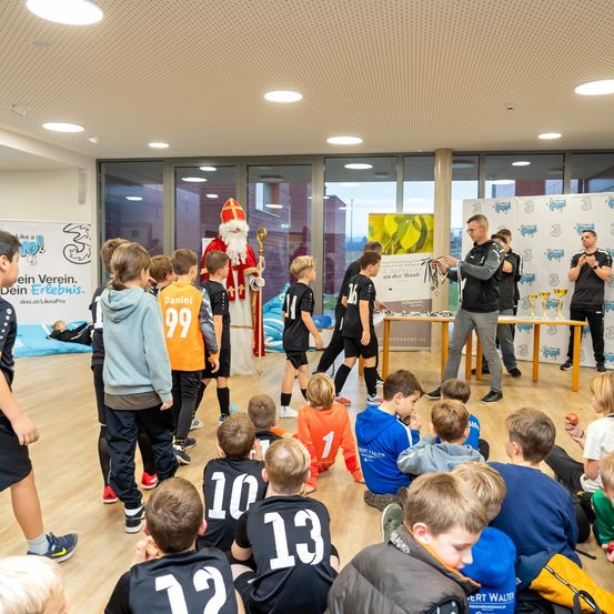 Eine Gruppe von Kindern in Trikots versammelt sich um einen Mann in einer Weihnachtsmann-Verkleidung. Der Mann verteilt Medaillen an die Kinder. Sie stehen und sitzen auf dem Boden. Es gibt Pokale und ein Banner auf einem Tisch. Daneben steht eine Person, und eine Glaswand ist dahinter.