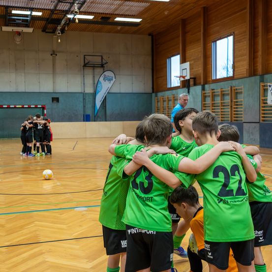 Eine Gruppe von Kindern in grünen Trikots ist auf einem Basketballplatz versammelt. Die Kinder haben Nummern auf ihren Trikots. Ein Mann in einem blauen Shirt steht hinter ihnen. In der Ferne liegt ein Fußball auf dem Boden.