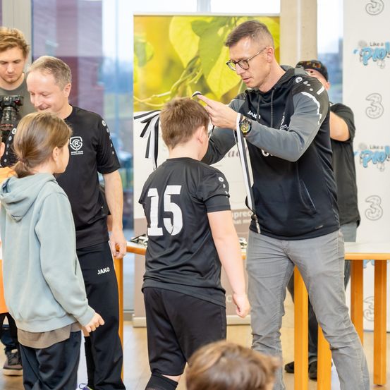 Ein Mann mit Brille befestigt eine Schleife auf dem schwarzen Trikot eines Jungen, Nummer 15, während ein anderer Mann in einem schwarzen Shirt zusieht. Ein Mädchen in einer grauen Kapuzenjacke beobachtet. Im Hintergrund befindet sich ein Banner mit einem Logo.