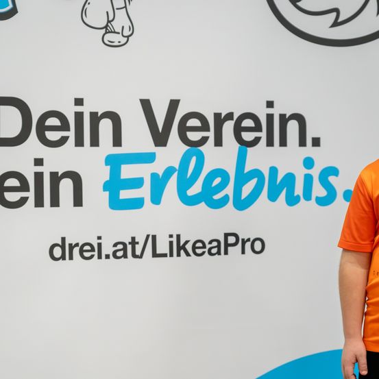 Eine Person in einem orangefarbenen Shirt steht vor einer weißen Wand mit einem blau-weißen Design. Es steht 'Dein Verein. Dein Erlebnis' in großen Buchstaben. Auf der Wand befindet sich auch ein Cartoon-Charakter.