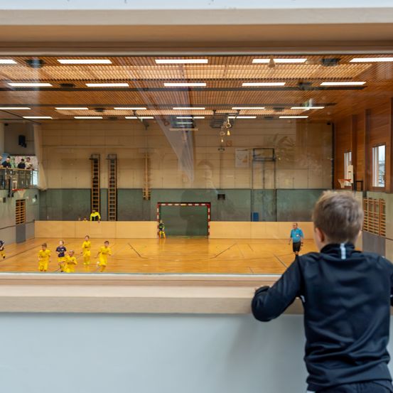 Ein junger Junge beobachtet Kinder, die Basketball in einer Turnhalle spielen, durch ein Glasfenster. Die Spieler tragen gelbe Trikots.