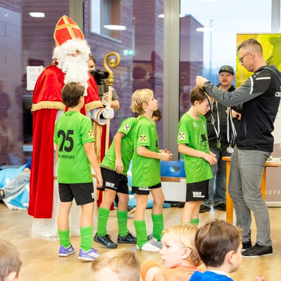 Eine Gruppe von Kindern in grünen Trikots steht neben einem Mann in einem Weihnachtsmannkostüm und einem Mann, der die Haare eines Jungen in Ordnung bringt.