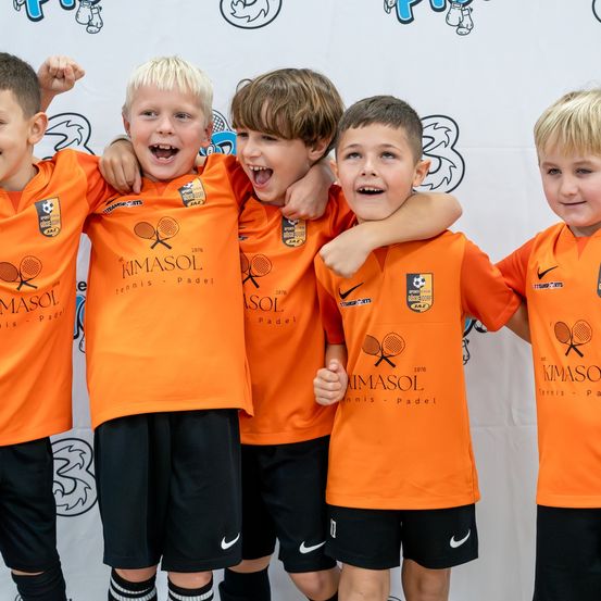 Eine Gruppe junger Jungen in orangefarbenen Sporttrikots mit dem Wort KIMSOL darauf. Sie stehen eng beieinander und lächeln fröhlich.
