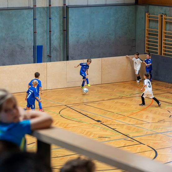 Mehrere junge Jungen spielen auf einem Hallensportplatz Fußball. Ein Junge tritt den Ball, während andere stehen und zusehen. Holzwände umgeben den Platz.