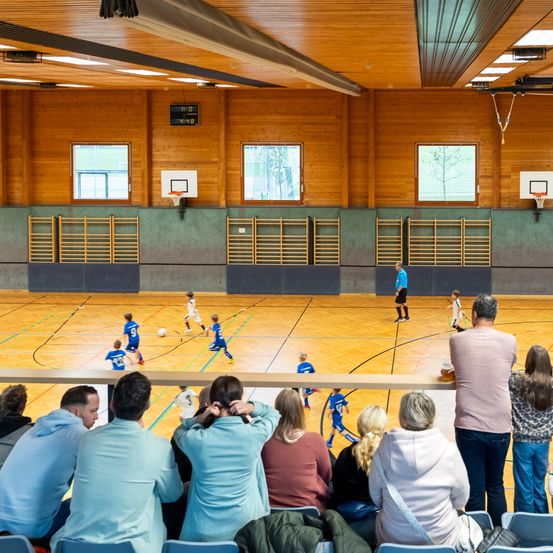 Eine Sporthalle mit Holzwänden und einem Basketballfeld. Mehrere Kinder spielen Fußball auf dem Platz, während einige Erwachsene von den Tribünen zusehen.