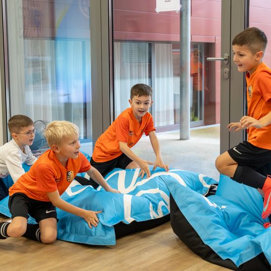 Vier Jungen spielen drinnen mit Sitzsäcken, zwei auf dem Boden und zwei stehend, alle tragen orangefarbene Shirts. Ein Junge hält einen Sitzsack, ein anderer steht in der Nähe einer Tür und ein weiterer in der Nähe eines Glasfensters.