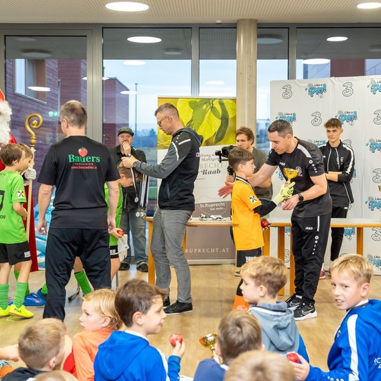 Eine Gruppe von Kindern und Erwachsenen in Sportkleidung versammelt sich in einem Saal. Ein Mann überreicht einem Kind eine Trophäe, während andere zusehen. Banner mit Logos hängen an der Wand. Der Weihnachtsmann steht an der Seite.