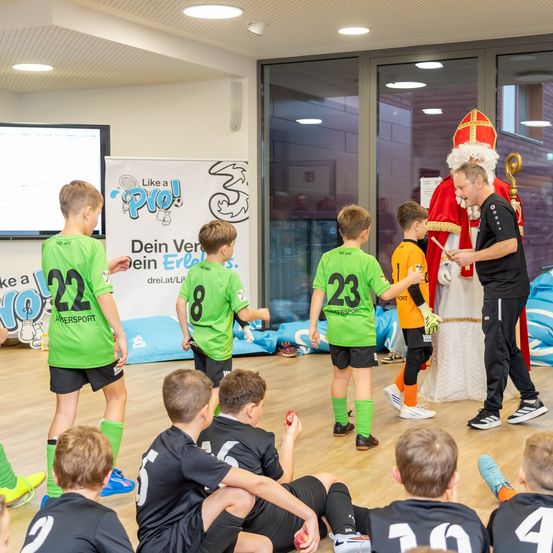 Eine Gruppe von Jungen in Sporttrikots nimmt an einer Veranstaltung mit einem Mann als Weihnachtsmann teil. Sie befinden sich in einem Raum mit einer Projektionsleinwand, einem Banner und Glastüren.