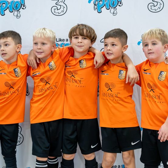Fünf Jungen in orangefarbenen Trikots mit Tennisschlägern und KIMASOL darauf stehen nebeneinander. Der Hintergrund ist weiß mit verschiedenen Sportlogos.