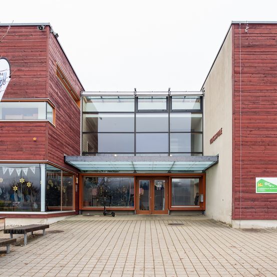 Ein modernes Gebäude mit roter Fassade und Glasfenstern. Es hat einen gläsernen Eingang mit Vordach und einen kleinen Roller davor.