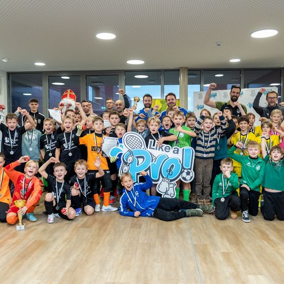 Eine Gruppe junger Athleten posiert für ein Foto mit Medaillen, Trophäen und einem Schild, das 'Wie ein Profi!' besagt. Sie befinden sich in einem Raum mit großen Glasfenstern.