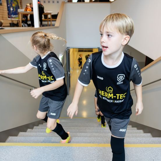 Zwei Kinder in identischen Sporttrikots laufen eine Treppe hinauf, eines in gelben Schuhen und das andere in grünen. Dahinter zeigt ein verschwommener Hintergrund einen Flur mit einem Geländer.