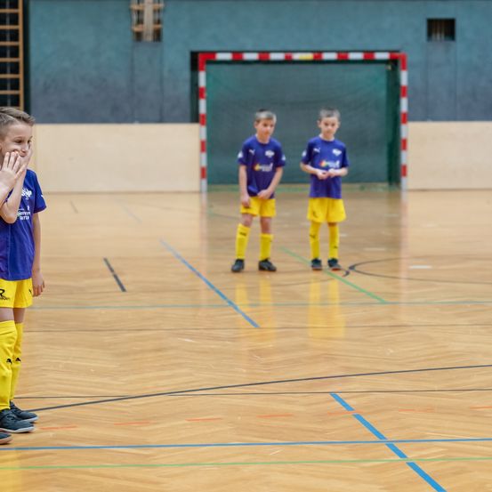 Eine Gruppe junger Jungen in blauen und gelben Trikots steht auf einem Spielfeld und wartet möglicherweise auf ein Spiel. Ein Junge berührt seinen Mund, während zwei andere bei der Torwartecke stehen.