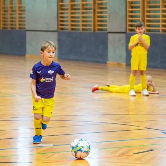 Drei Kinder sind auf einem Spielfeld. Ein Junge läuft mit einem Fußball. Ein anderer Junge beobachtet ihn. Ein Mädchen liegt auf dem Boden.