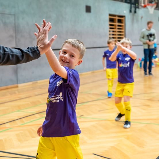 Mehrere Jungen spielen Fußball auf einem Spielfeld. Ein Junge im Vordergrund bekommt eine High-Five. Im Hintergrund filmt ein Mann mit einer Kamera.