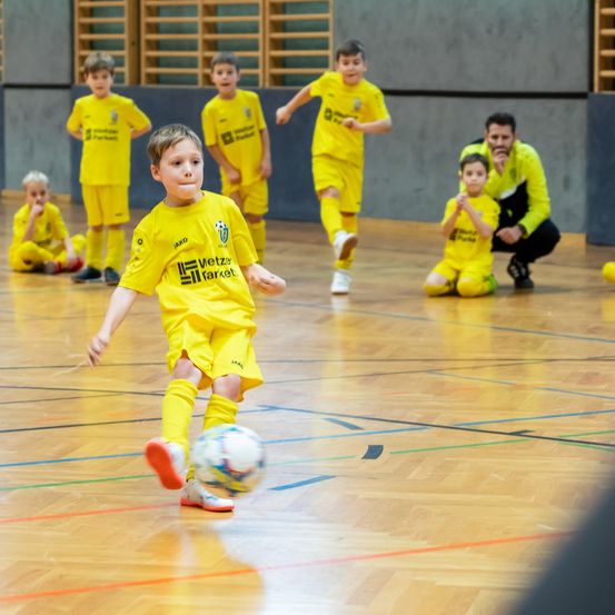 Kinder in gelben Fußballtrikots spielen auf einem Spielfeld. Ein Junge tritt einen Fußball. Hinter ihm beobachtet ein Mann. Andere Kinder sitzen an den Seitenlinien.