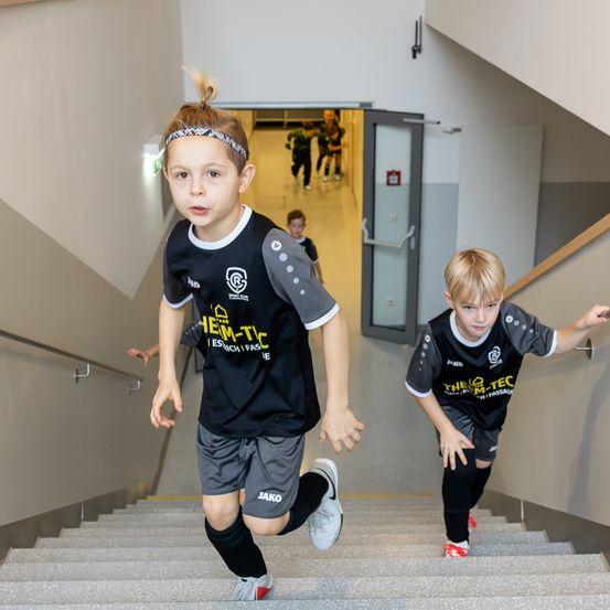 Zwei Jungen rennen die Treppe hinauf. Sie tragen schwarze und graue Sporttrikots. Ein Feuerlöscher ist an der Wand montiert.